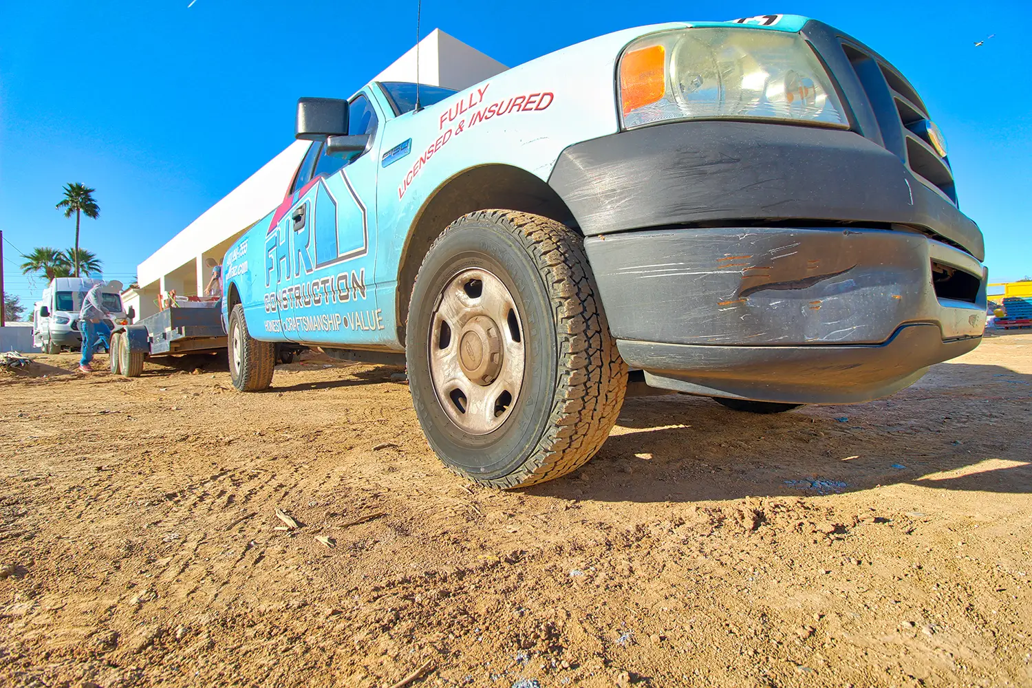 A blue pickup truck with a company logo sits on a dirt lot, its front bumper scratched. It showcases Fully Licensed & Insured and services like home renovation. A palm tree and a white building stand under a clear blue sky. Another truck, possibly from general contractor Scottsdale, is to the left.