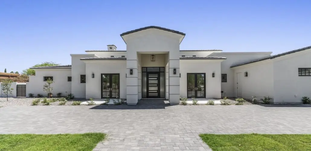 A modern white house with a flat roof, crafted by one of the best home remodeling contractors near me, features a prominent central entrance with black-framed glass doors and windows. Gray stone pavers form the driveway, and small plants line the front. The clear blue sky enhances its architectural lines.