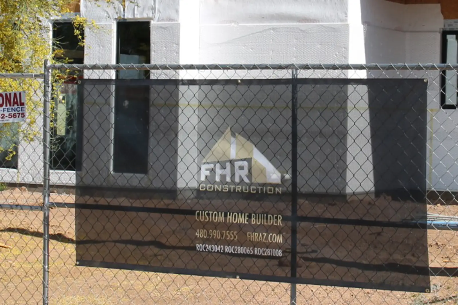 A construction site is enclosed by a chain-link fence with a black sign that reads FHR Construction in white and gold letters. This Custom Home Builder, known for top-tier home renovation, displays contact info as a partially built house stands visible in the background.