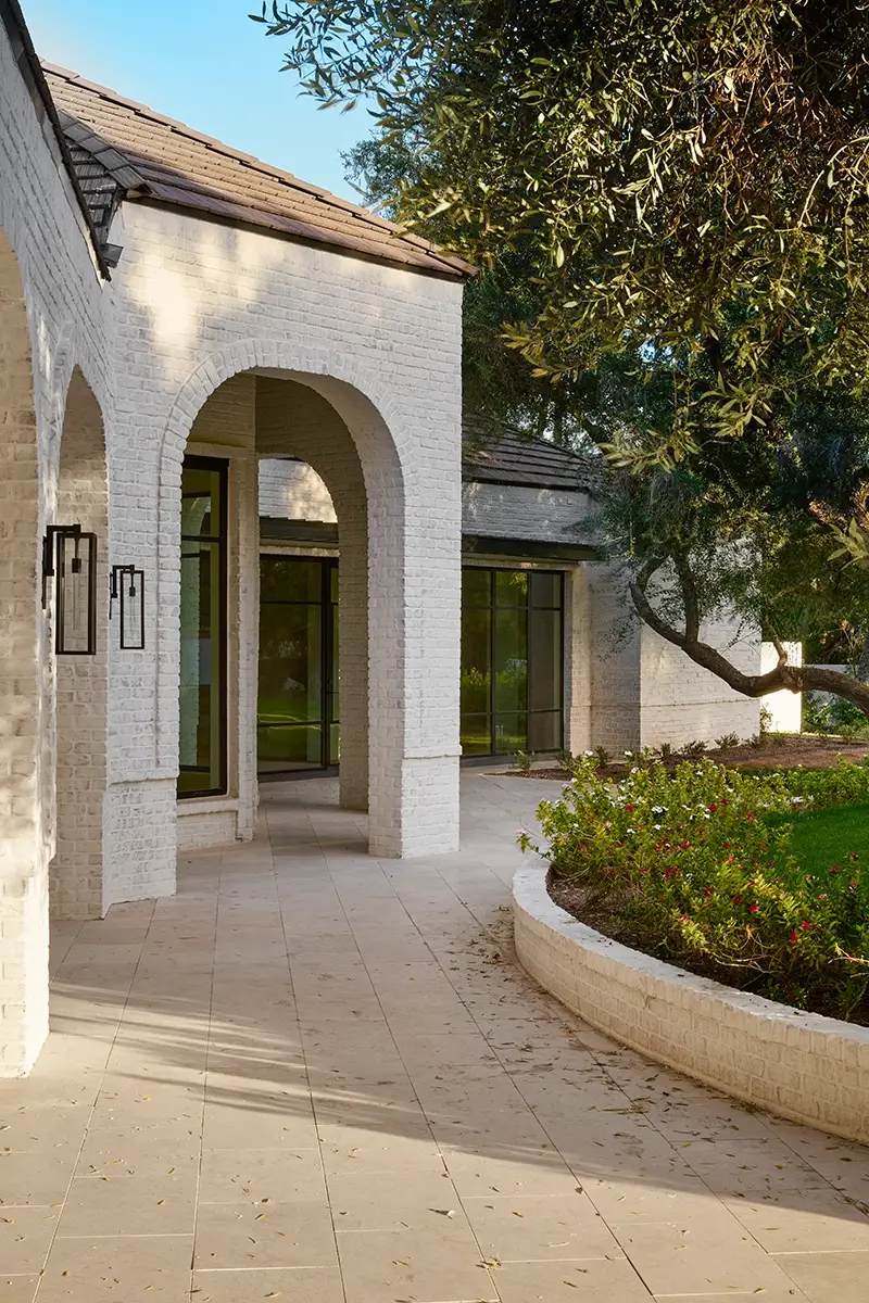 Elegant modern house exterior featuring white brick arches and a shaded front porch. Large windows line the walls, reflecting the lush greenery of the garden. A curving pathway with flowerbeds leads to the entrance, hinting at expert kitchen remodeling inside that blends style with functionality.