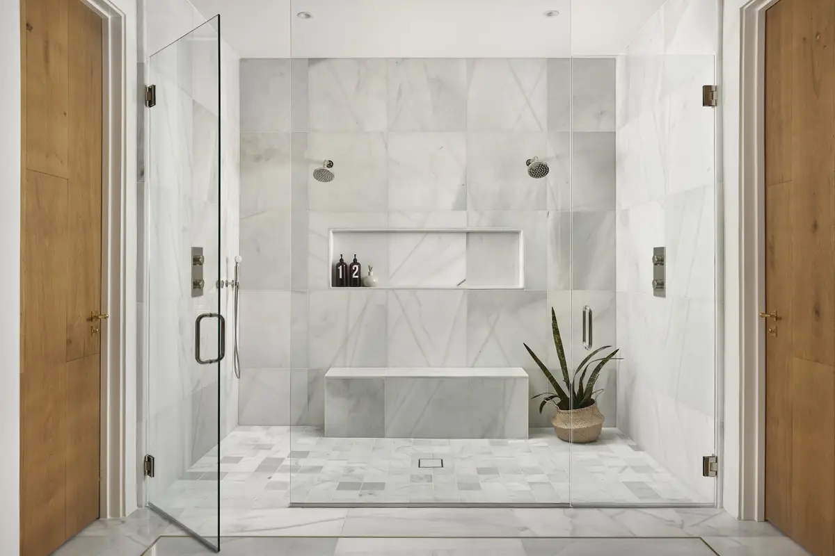 Experience a modern bathroom renovation with elegant marble tiles on walls and floor. The glass shower enclosure features a built-in bench and a recessed shelf for soap bottles. A potted plant adds charm, while wooden doors and recessed ceiling lights complete this stylish transformation.