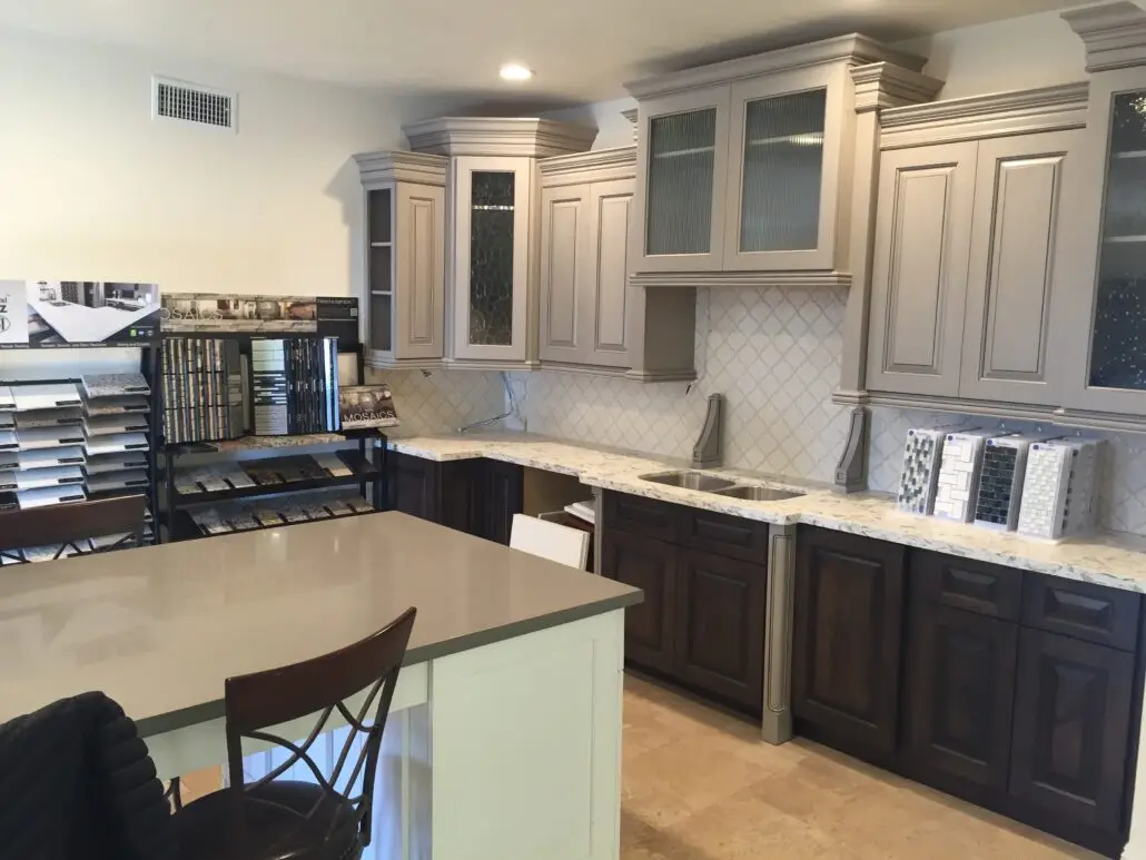 A showroom features a kitchen display with two-tone cabinets: light gray uppers with glass panels and dark wood lowers. Marble-patterned countertops and a tile-patterned backsplash are visible. Sample boards and catalogs are arranged on counters and racks nearby.