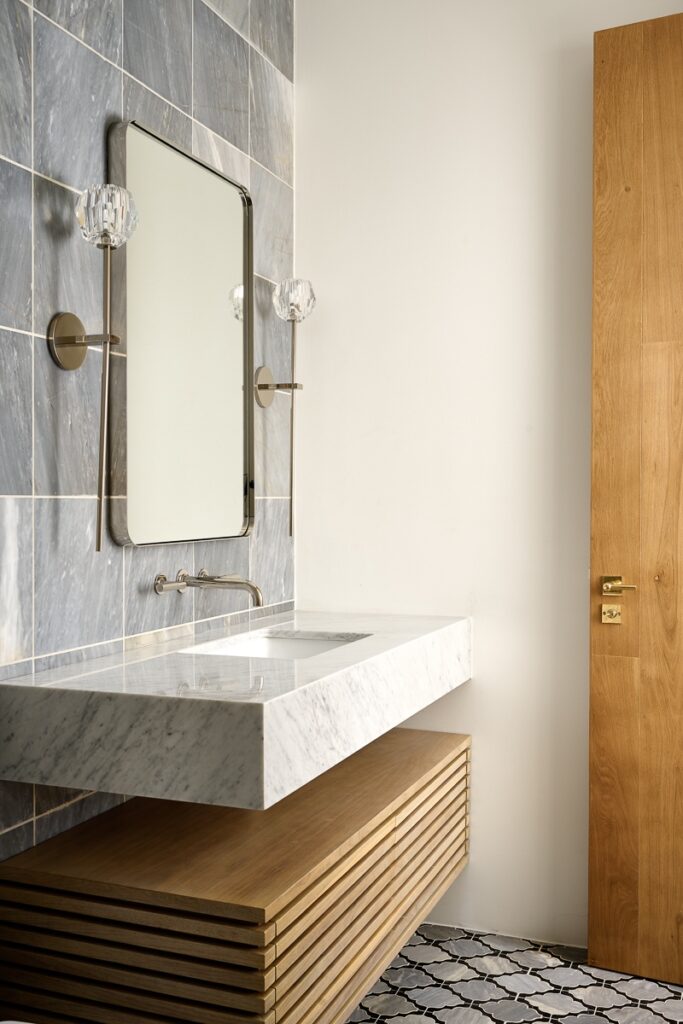 Modern bathroom makeover featuring a marble sink, wall-mounted faucet, rectangular mirror, two wall sconces, gray tile backsplash, wooden vanity, and patterned floor tiles.