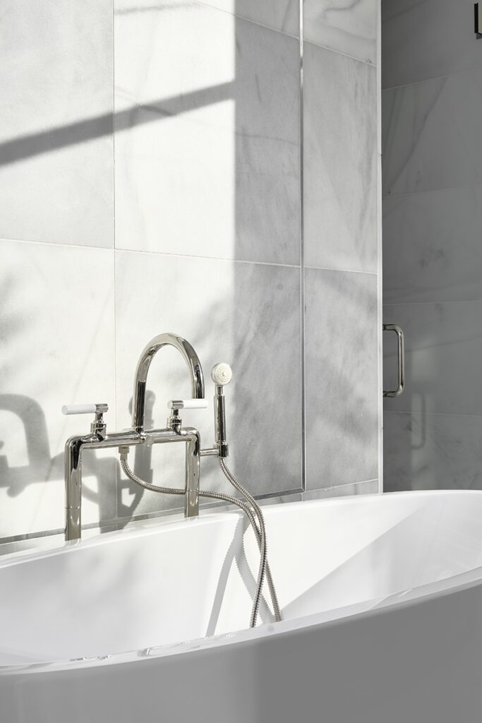 A modern bathtub with chrome fixtures and a hand shower sits against a wall of light gray marble tiles in this sunlit bathroom, showcasing elegant style perfect for any Bathroom Renovations.