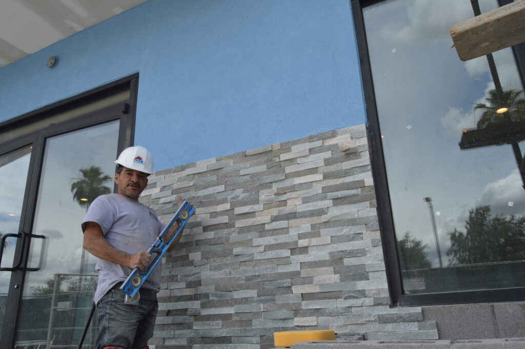 A construction worker in a hard hat levels a newly installed stone veneer wall on the exterior of a commercial remodeling project with blue siding and large windows.