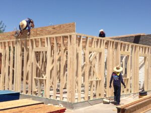 Three construction workers wearing hats are building a wooden frame structure under a clear sky, working together on a commercial remodeling project. One worker stands on the roof while two others assist from the ground.