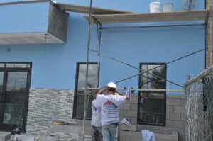 Two construction workers in hard hats stand near scaffolding beside a blue building with tiled and brick walls—a scene typical of commercial remodeling; buckets rest on the wooden platform above.