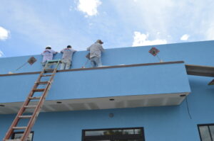 Three workers on a blue building’s roof install equipment as part of a commercial remodeling project, with a ladder leaning against the wall under a partly cloudy sky.