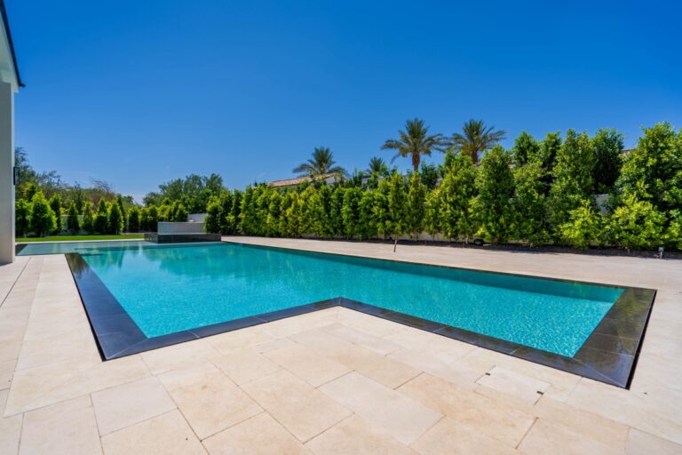 Rectangular outdoor swimming pool with clear blue water, surrounded by beige stone tiles, green hedges, and palm trees—perfect for stylish outdoor living under a clear sky.