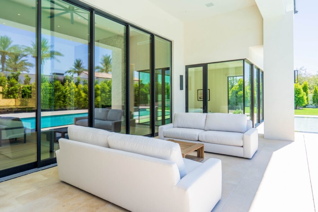 modern outdoor patio designed for outdoor living, featuring two white sofas and a wooden coffee table, positioned next to large glass doors overlooking a pool and landscaped garden.