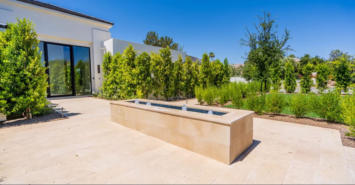 Rectangular stone fountain in a tiled patio offers serene outdoor living, surrounded by green shrubs and trees next to a modern building with large glass doors.