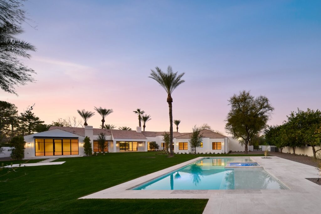 Modern single-story house with large windows, palm trees, and a rectangular swimming pool creates an inviting outdoor living space in a spacious, well-manicured backyard at sunset.