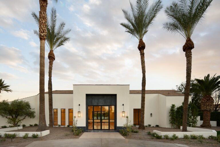 Modern single-story white house with large front windows, double glass doors, minimal landscaping, tall palm trees, and inviting outdoor living spaces set against a partly cloudy sky.