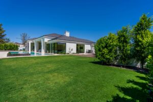 Modern single-story house with large windows, covered patio for seamless outdoor living, swimming pool, and well-maintained backyard lawn under a clear blue sky.