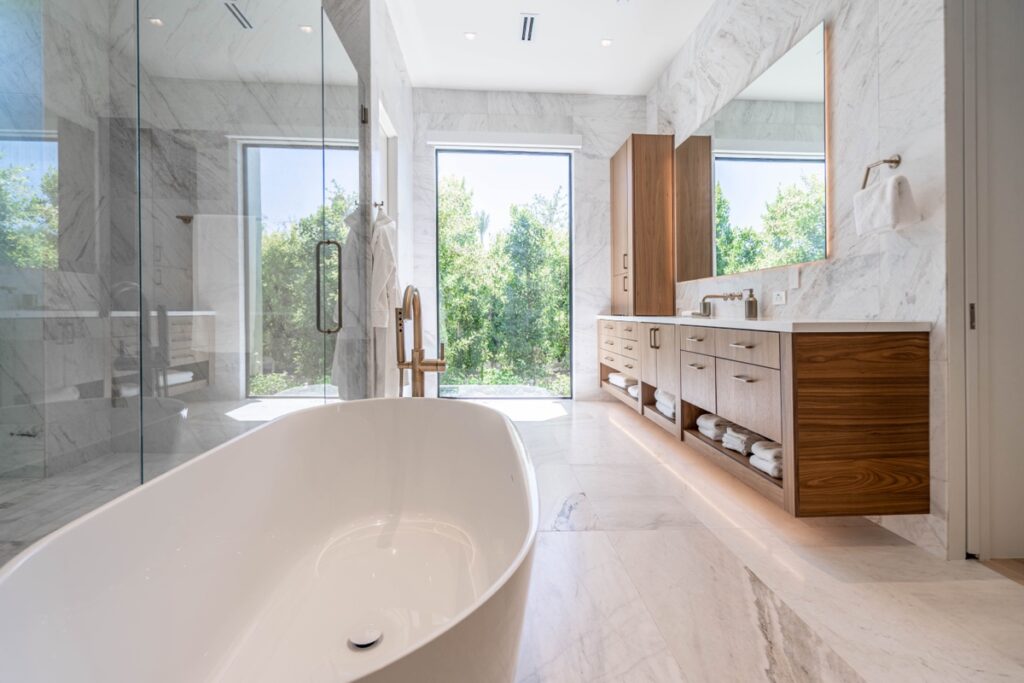 Modern bathroom with a freestanding bathtub, glass shower, wooden vanity, large mirrors, marble floors and walls—an ideal space for bathroom renovations overlooking lush greenery through large windows.