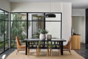 Modern dining room with a black table, six mixed chairs, two large vases with foliage, floor-to-ceiling windows, and a pendant light above on a neutral woven rug.