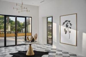 Modern entryway with large black-framed glass doors, a geometric chandelier, a round table with abstract sculptures, and a black-and-white botanical artwork on the wall.