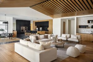 Open-plan living room with beige sofas, round poufs, wood flooring, and ceiling accents, seamlessly connected to a kitchen and dining area with black and wood cabinetry.