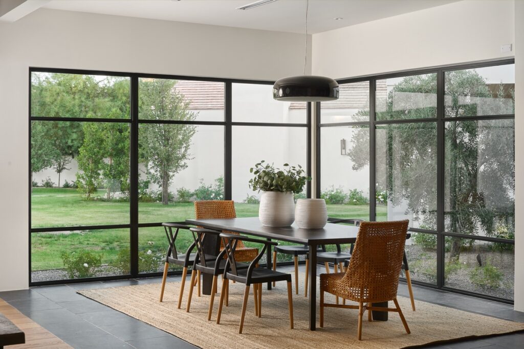 Modern dining area with a black table, mixed chairs, two vases with greenery, large windows, and a view of a garden outside.