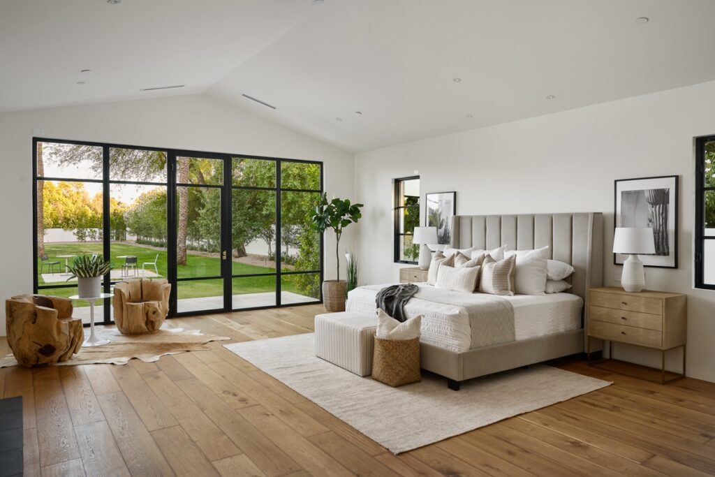 Modern bedroom with a beige upholstered bed, wooden side tables, large windows overlooking a garden, indoor plant, and light wood flooring.