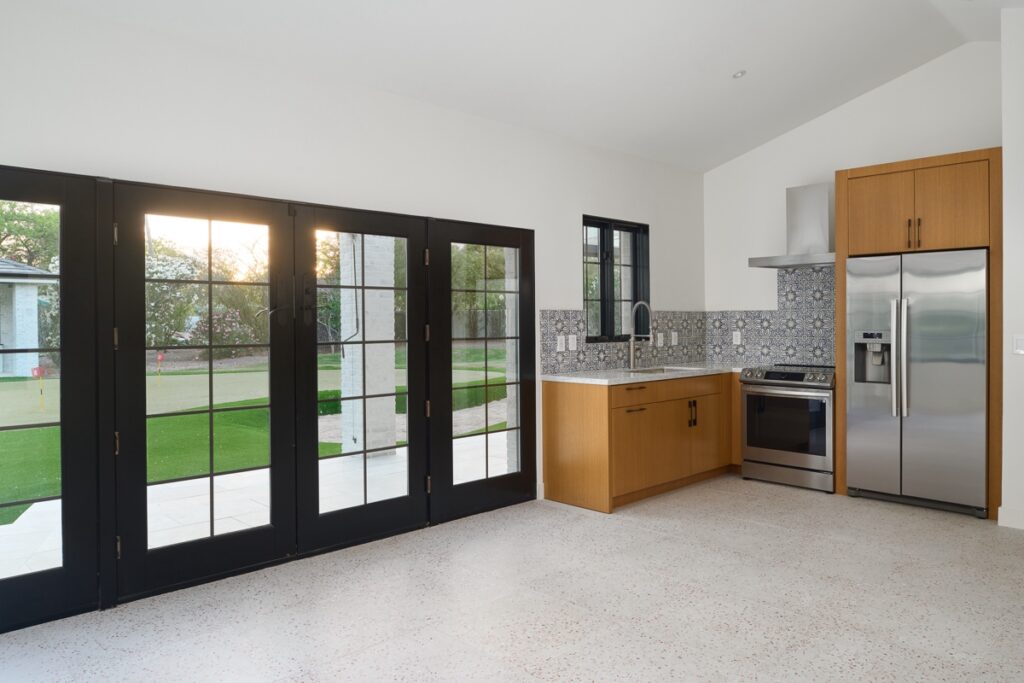Modern kitchen with light wood cabinets, stainless steel appliances, patterned backsplash, and large black-framed glass doors overlooking a green lawn.