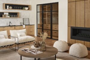 Modern living room with light wood cabinets, white cushioned chairs, a round coffee table, two poufs, a fireplace, and a glass wine storage room in the background.