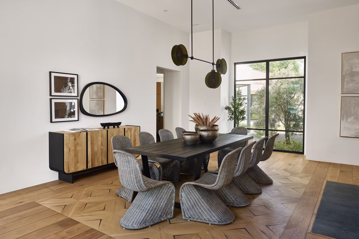 Modern dining room with a long black table, ten woven chairs, wooden sideboard, wall art, and large window letting in natural light.
