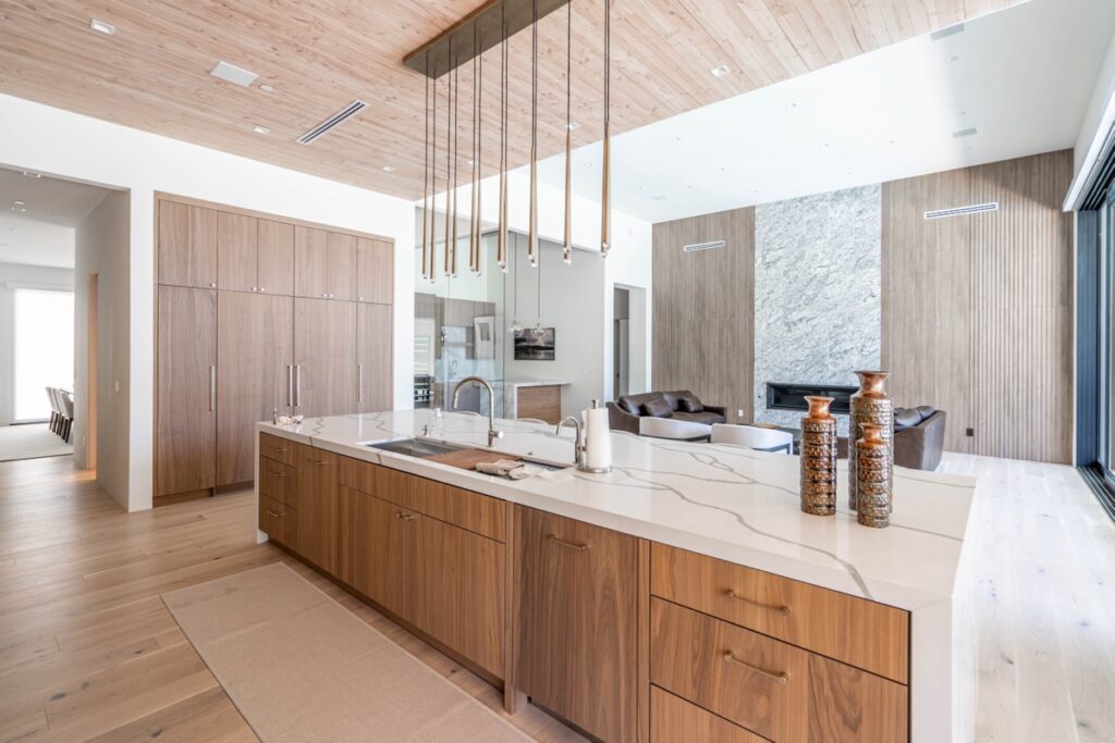 Modern kitchen remodeling features a large island, wooden cabinetry, stylish hanging light fixtures, and a spacious open floor plan connecting to a living area with a stone accent wall and fireplace.