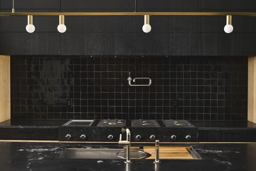 Modern kitchen remodel with black tiled backsplash, black countertops, built-in stovetop, stainless steel sink, and a gold light fixture with exposed bulbs above—perfect inspiration for your next kitchen renovation.
