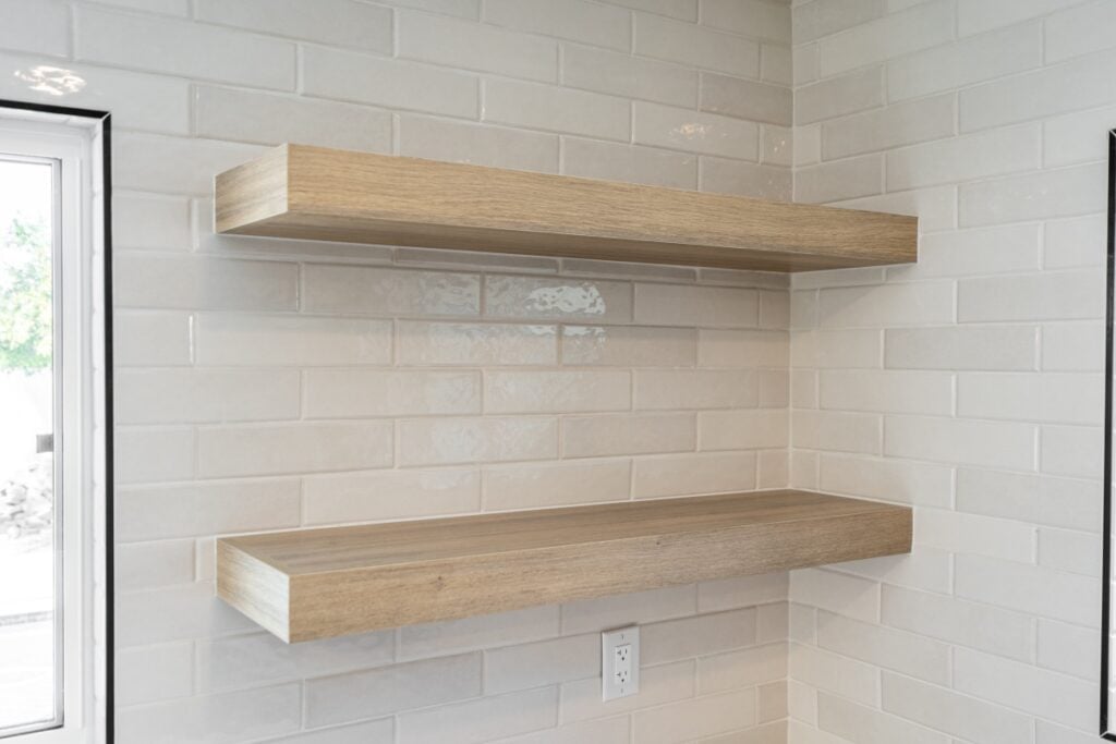 Two wooden floating shelves are mounted on a white tiled wall above an electrical outlet, near a window—perfect for showcasing your kitchen remodeling style.