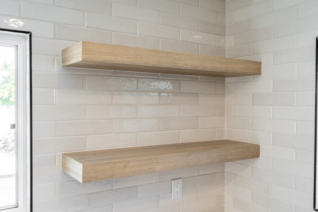 Kitchen Remodeling Two wooden floating shelves are mounted on a white tiled wall above an electrical outlet, near a window—perfect for showcasing your kitchen remodeling style.