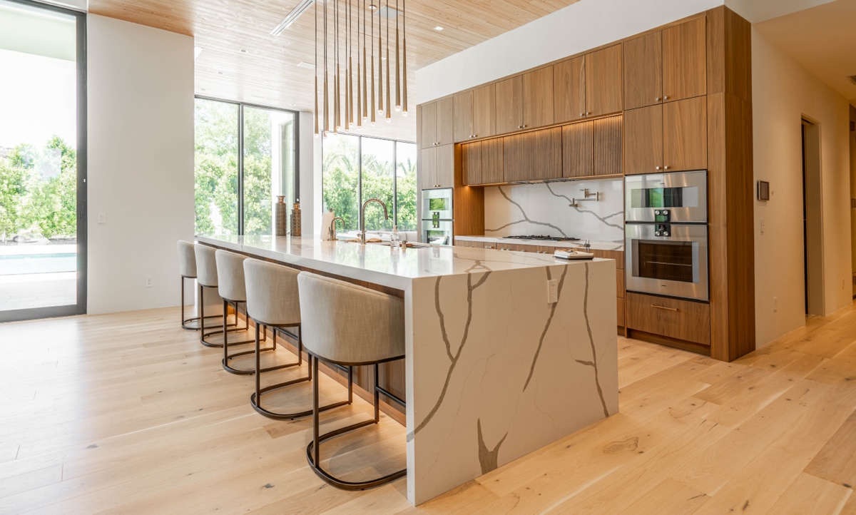Thank you Modern kitchen remodeling features light wood flooring, a spacious marble island with barstools, sleek wood cabinets, built-in appliances, and floor-to-ceiling windows overlooking lush greenery.