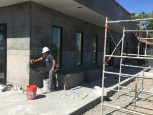 A construction worker wearing a hard hat applies material to a cinder block wall near scaffolding during a commercial remodeling project at a building site on a sunny day.
