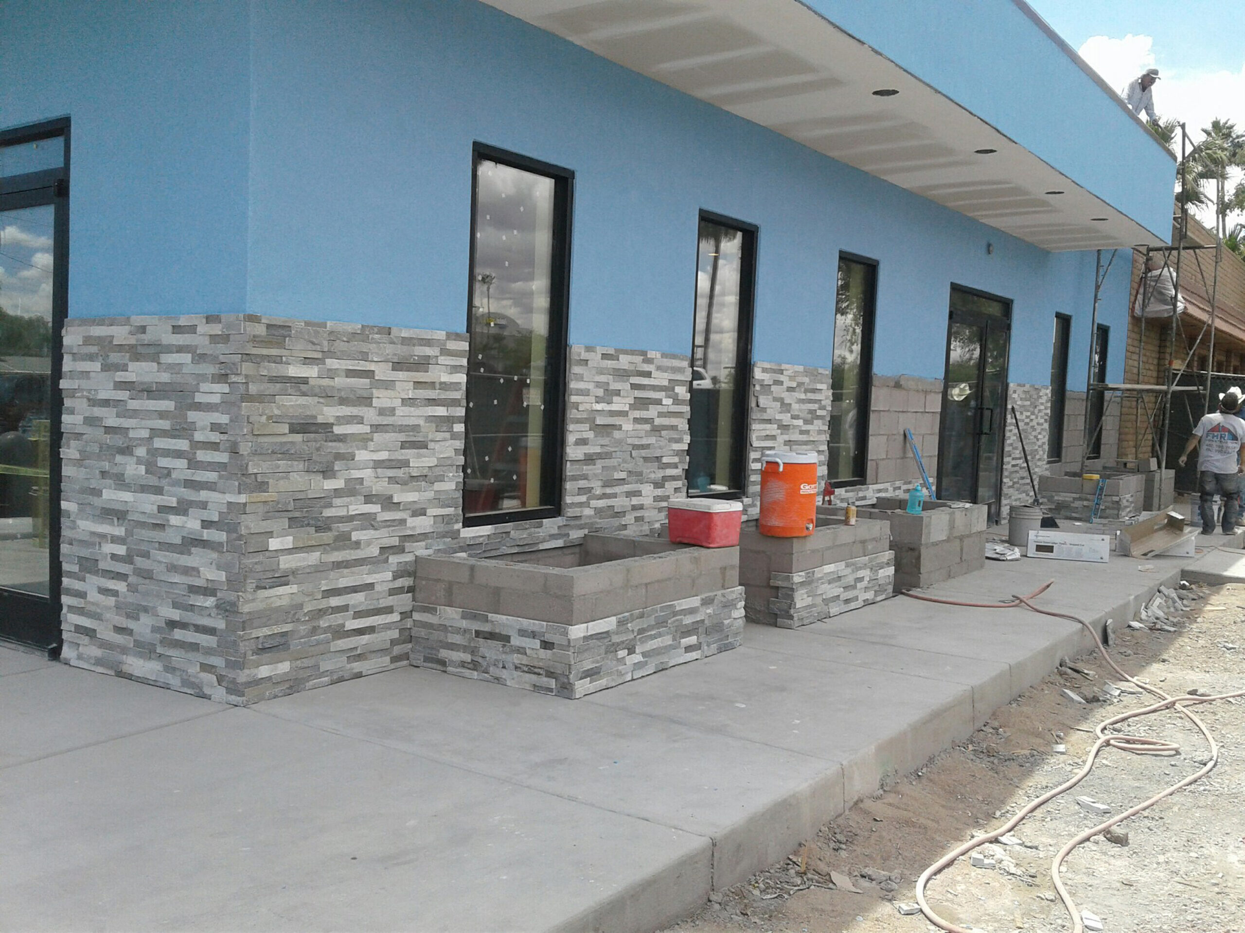 A building under construction undergoing commercial remodeling, featuring blue walls, stone veneer on the lower exterior, unfinished planters, and construction materials on the sidewalk.