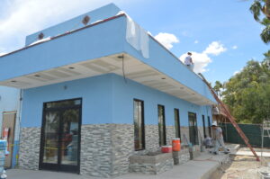 Workers are painting and renovating the exterior of a blue building with stone trim under a partly cloudy sky, showcasing expert commercial remodeling.