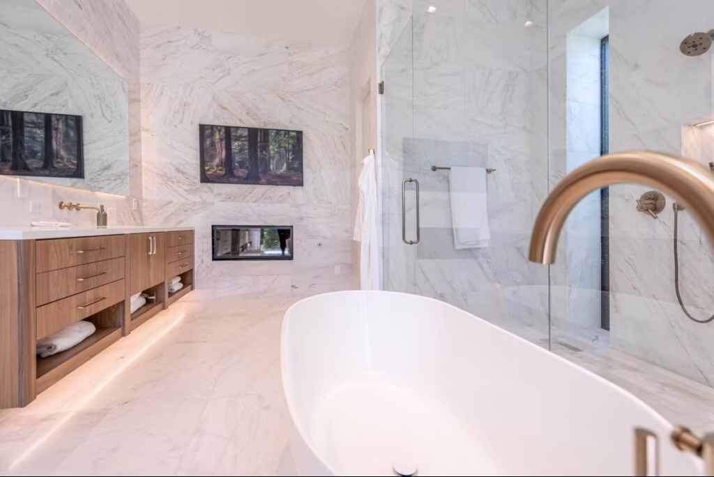 Experience a stunning bathroom makeover featuring a freestanding bathtub, marble walls and floors, wood cabinetry, glass shower, gold fixtures, and a wall-mounted TV above the fireplace.