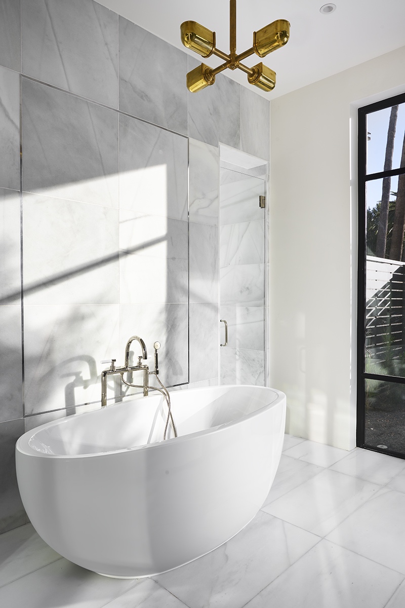 modern bathroom with a white freestanding bathtub, marble tile walls and floor, a gold ceiling light fixture, and large windows letting in natural light.