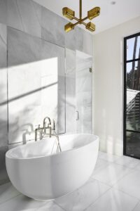Modern bathroom with a white freestanding bathtub, marble tile walls and floor, a gold ceiling light fixture, and large windows letting in natural light.