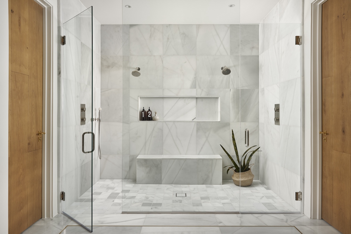 spacious marble shower with dual showerheads, a built in bench, two wooden doors, glass enclosure, and a potted plant on the floor.