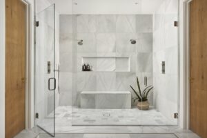 Spacious marble shower with dual showerheads, a built-in bench, two wooden doors, glass enclosure, and a potted plant on the floor.