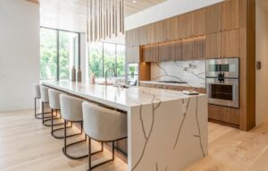 Modern kitchen with a large marble island, five upholstered bar stools, wood cabinetry, built-in ovens, and large windows providing natural light.