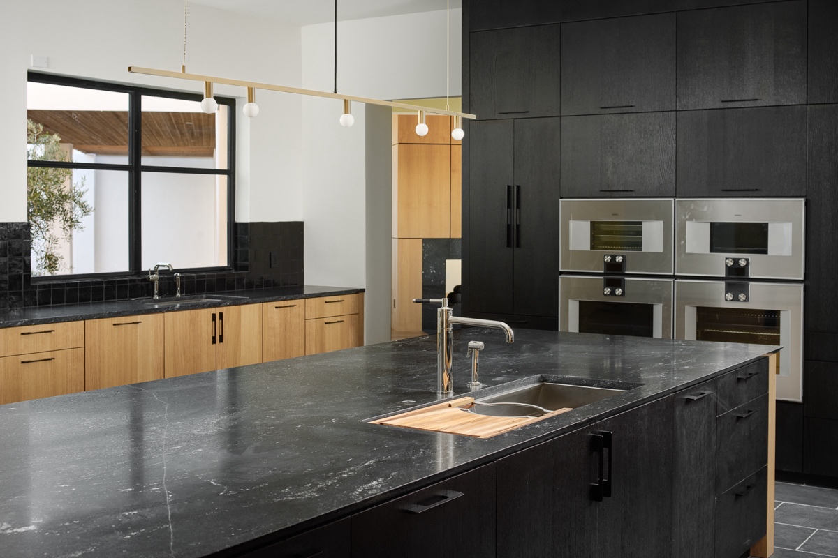 Modern kitchen with black countertops and cabinets, light wood lower cabinets, built-in ovens, large island with sink, and a minimalist pendant light fixture.