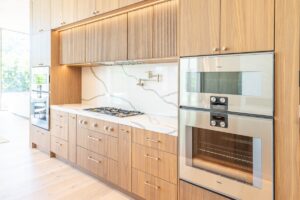 Modern kitchen with light wood cabinets, built-in stainless steel double ovens, a gas cooktop, marble backsplash, and under-cabinet lighting.