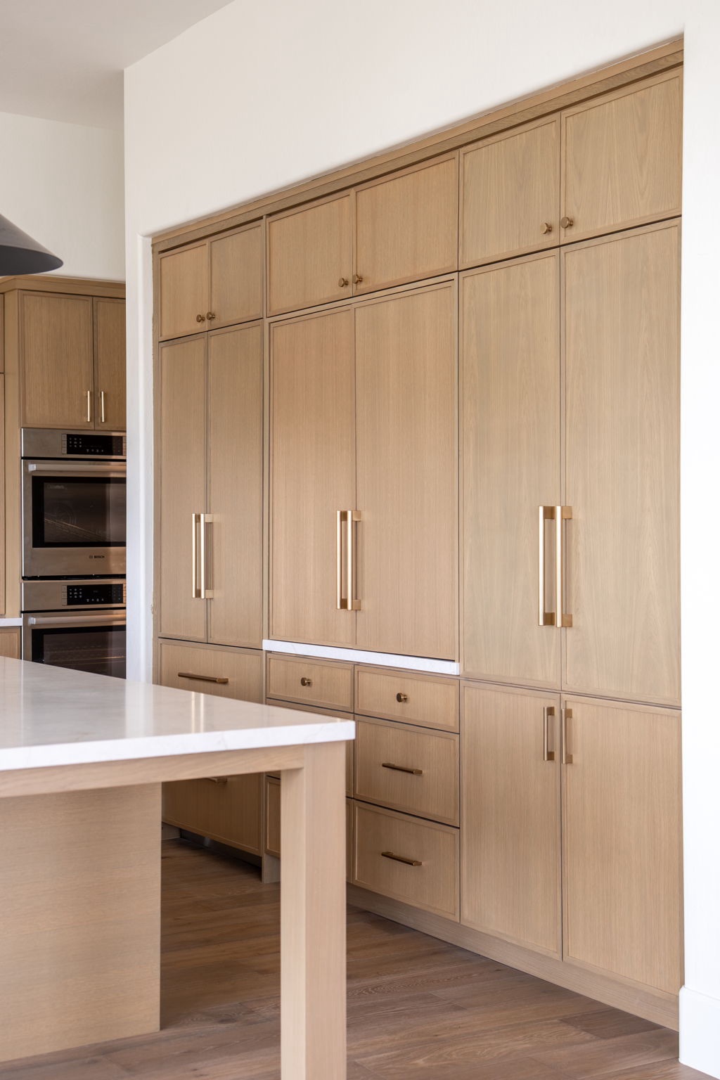 light wood kitchen cabinets with gold handles, a built in oven, and a matching island with a white countertop are shown in a modern, minimalistic kitchen.
