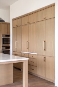 Light wood kitchen cabinets with gold handles, a built-in oven, and a matching island with a white countertop are shown in a modern, minimalistic kitchen.