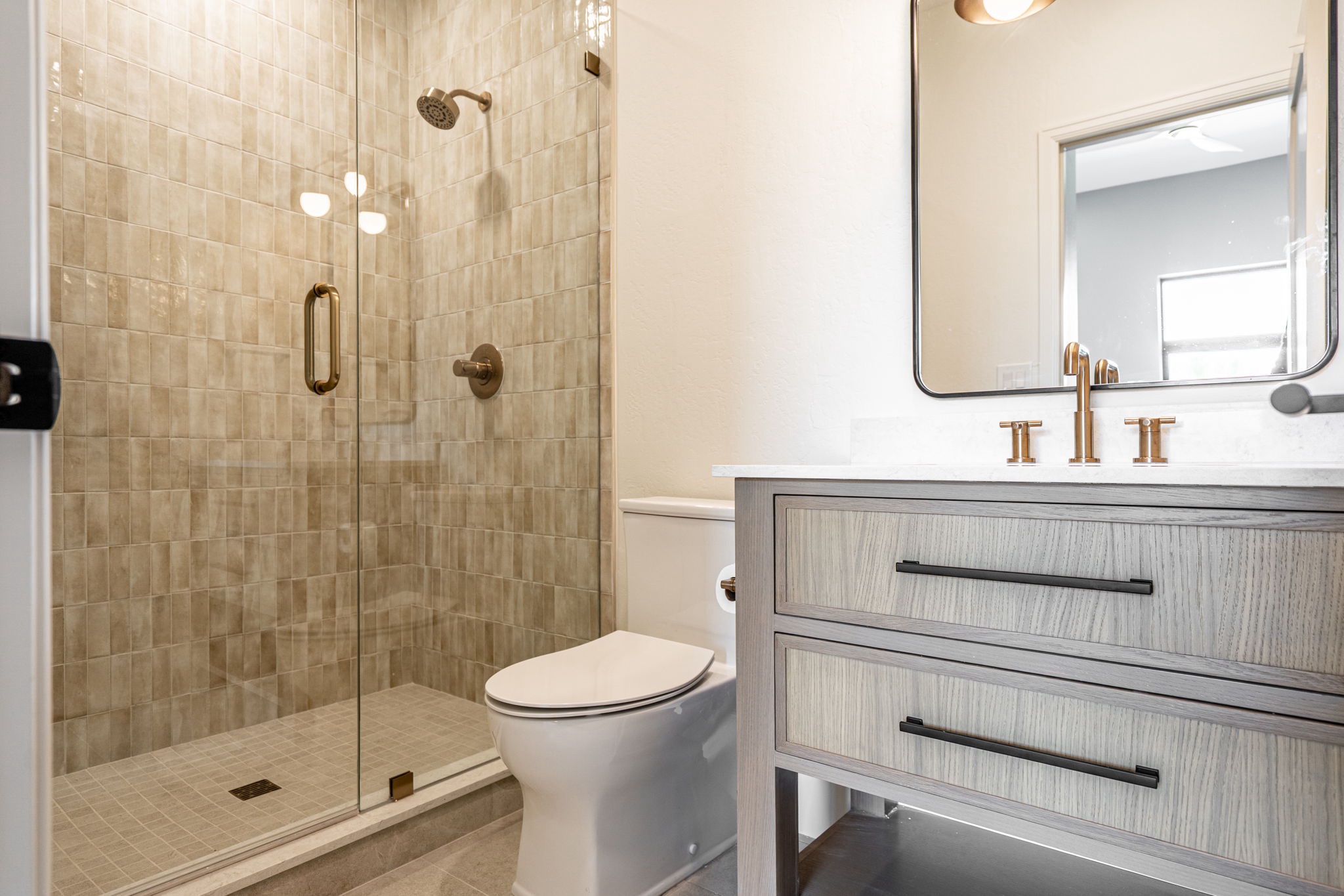 Modern bathroom with a glass-enclosed shower, tiled walls, a white toilet, and a vanity with a white countertop and bronze fixtures.