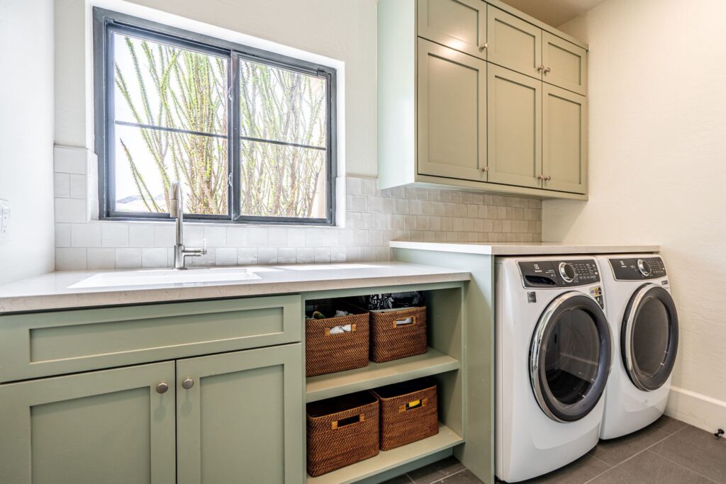 Laundry room with green cabinets, built-in sink, front-loading washer and dryer, and open shelves holding woven baskets under a window.