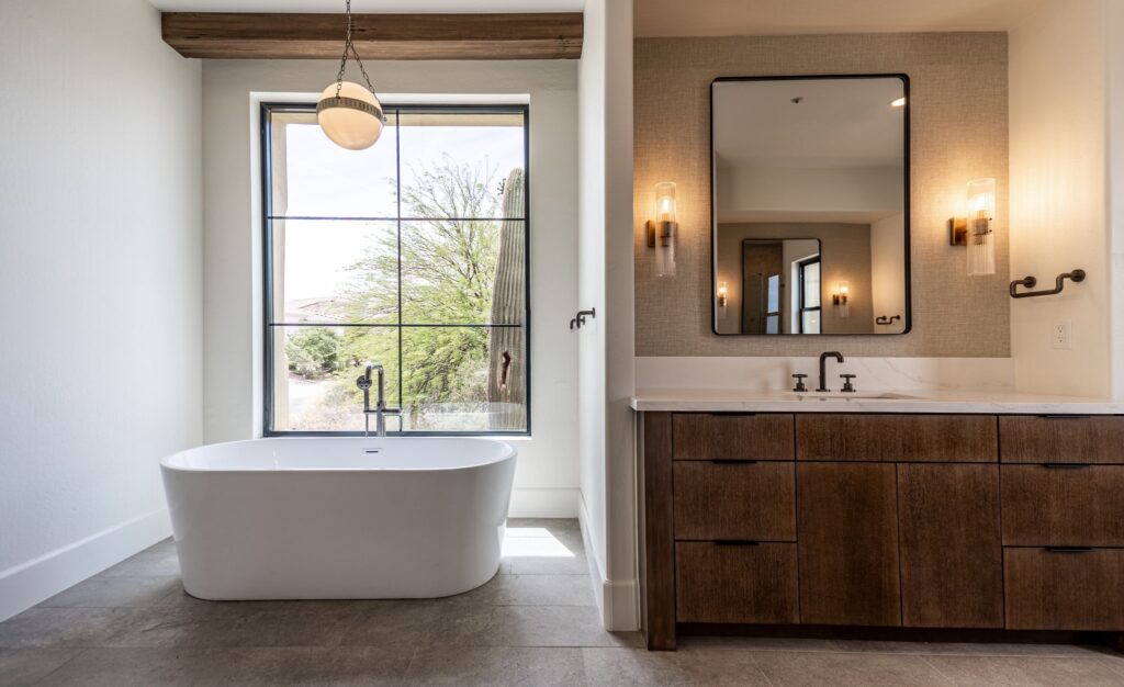 Modern bathroom with a freestanding bathtub near a large window, wooden vanity with double sinks, wall sconces, and a rectangular mirror.