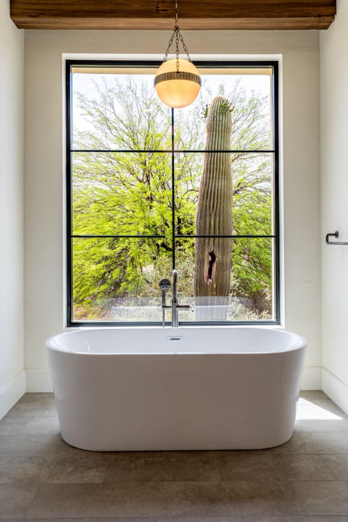 a modern white bathtub is positioned in front of a large window with black grid frames, showcasing a view of a tall cactus and green desert foliage outside.