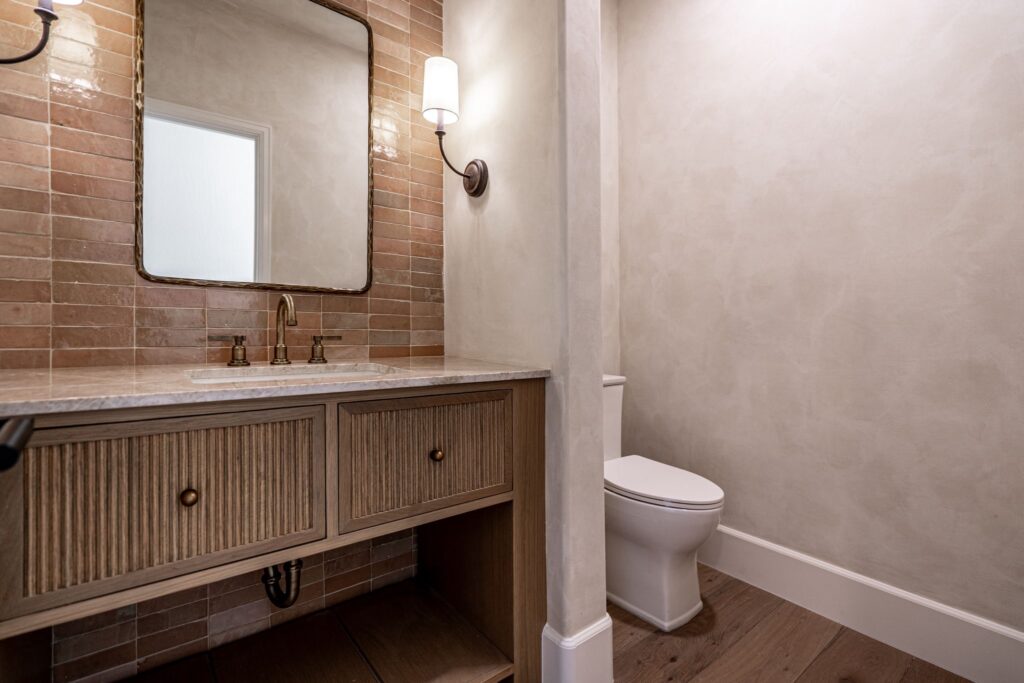 A bathroom with a wooden vanity, marble countertop, wall-mounted mirror, light fixture, and a separate area with a toilet and textured walls.